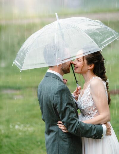 Ein Brautpaar steht eng umschlungen unter einem transparenten Regenschirm im Regen und berührt liebevoll mit den Nasen einander.