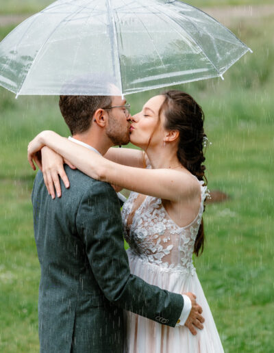 Das Brautpaar küsst sich innig unter einem durchsichtigen Regenschirm im Regen, die Braut umfasst den Bräutigam liebevoll.