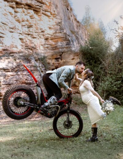 Ein sportlicher Bräutigam küsst seine schwangere Braut, während er auf einem Motorrad balanciert – ein Moment voller Charakter, eingefangen in echter Hochzeitsfotografie Lahr mit Persönlichkeit.