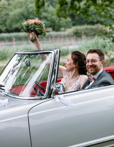 Fröhliches Brautpaar im Oldtimer – die Braut jubelt mit erhobenem Blumenstrauß. Helene Karsten Hochzeitsfotografie fängt pure Lebensfreude ein.