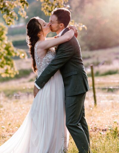 Kuss im goldenen Gegenlicht: Ein verliebtes Brautpaar umarmt sich im Abendlicht einer Sommerhochzeit – romantische Hochzeitsfotografie auf einer Wiese, die Emotionen sichtbar macht.