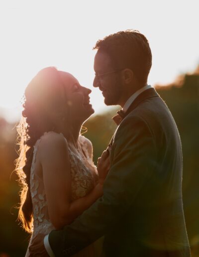 Eine Braut lacht voller Liebe in Richtung ihres Bräutigams, die Sonne leuchtet warm hinter ihnen. Eine emotionale Momentaufnahme einer freien Trauung im Sonnenuntergang.