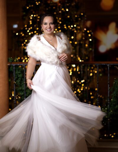 Elegante Braut in winterlichem Hochzeitskleid mit Kunstpelzstola vor funkelnden Weihnachtslichtern – ein festliches Wedding Shooting mit glamouröser Note.