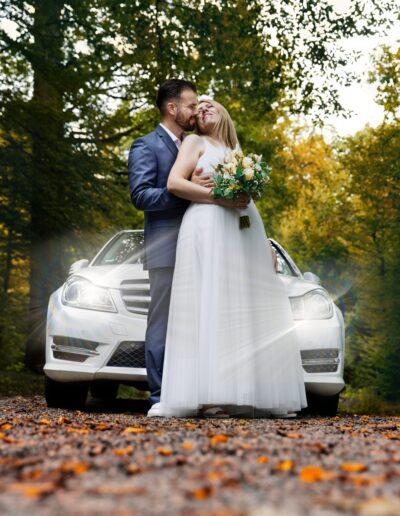 Innige Umarmung vor dem Hochzeitsauto – das Brautpaar genießt einen romantischen Moment unter herbstlich goldenen Baumkronen.