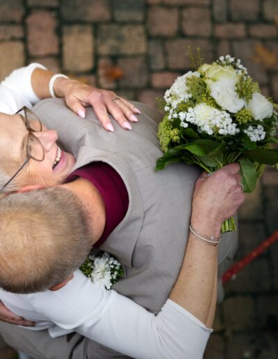 Eine ältere Braut lacht herzlich in der Umarmung ihres Partners – von oben eingefangen, mit Brautstrauß in der Hand. Hochzeit im Alter, echte Emotionen, Brautpaarumarmung, Vintage-Stimmung, natürliches Paarshooting.