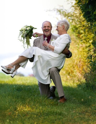 Der Bräutigam trägt seine Braut lachend durch eine Wiese – beide strahlen pures Glück und Leichtigkeit aus. Lebensfreude, Hochzeit im Grünen, Braut tragen, Outdoor-Hochzeit, Senior Wedding.