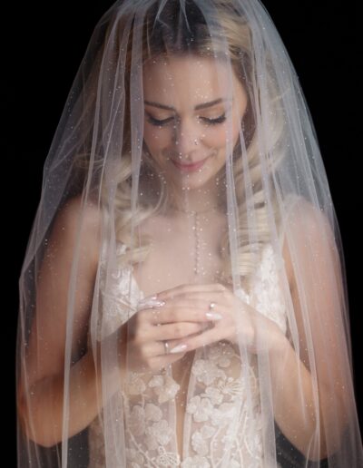 Zarte Nahaufnahme einer Braut im Spitzenkleid, die verträumt unter einem mit feinem Glitzer versehenen Brautschleier lächelt, während sie ihre Hände sanft hält.