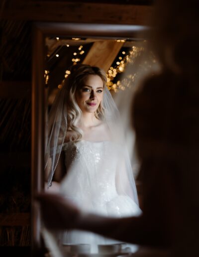 Elegantes Porträt einer Braut, die mit sanftem Lächeln in den Spiegel schaut, umgeben von goldenen Lichterketten in einer rustikalen Hochzeitslocation.
