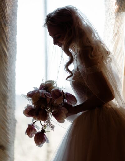 Künstlerische Silhouette der Braut im Spitzenkleid, die unter ihrem Schleier einen Strauß aus Magnolien und Rosen hält, sanft vom Tageslicht umspielt.