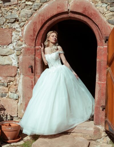 Eine strahlende Braut im voluminösen Prinzessinnen-Hochzeitskleid lehnt elegant im Torbogen eines alten Steingebäudes.