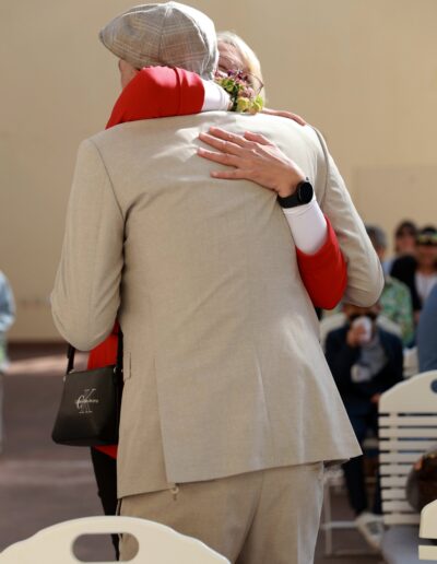 Der Bräutigam umarmt herzlich eine Frau in rotem Kleid und Blütenarmband, während im Hintergrund Gäste die emotionale Szene beobachten.