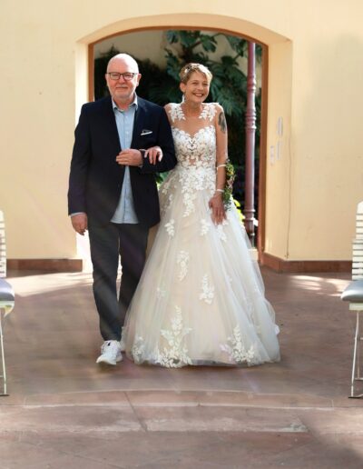 Strahlend schreitet die Braut im floralen Spitzenkleid mit einem älteren Herren durch den Torbogen, bereit für die Trauung.
