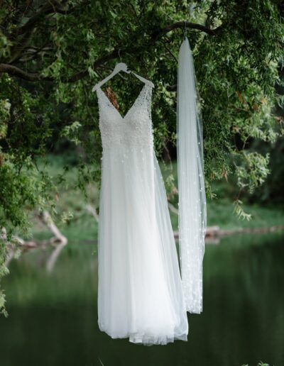 Ein zartes, besticktes Brautkleid und ein Schleier hängen romantisch an einem Ast am Seeufer.