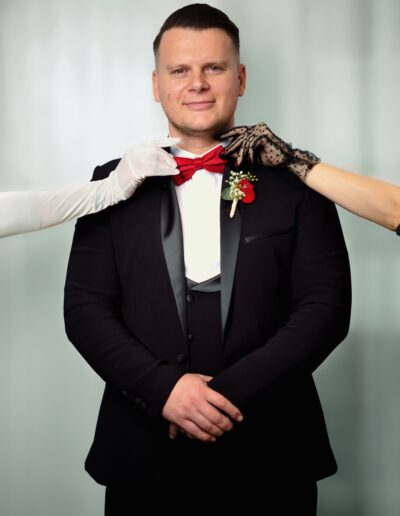 Ein Bräutigam im eleganten Smoking mit roter Fliege steht gelassen da, während mehrere Hände an seinem Outfit Details richten – stilvolle Hochzeitsvorbereitung.