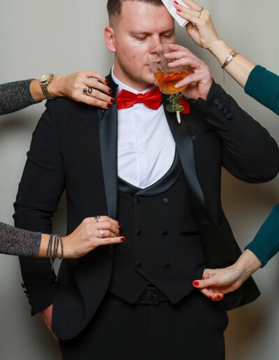 Der Bräutigam genießt einen Drink, während mehrere Hände ihm die Haare richten, sein Sakko zurechtzupfen und Details korrigieren – humorvolle Hochzeitsreportage.