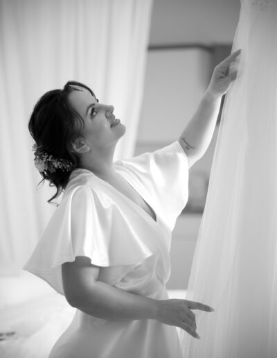 In zartem Schwarz-Weiß schaut die Braut liebevoll auf ihr Hochzeitskleid, das sie gleich anziehen wird – ein Moment der Vorfreude und Emotion.