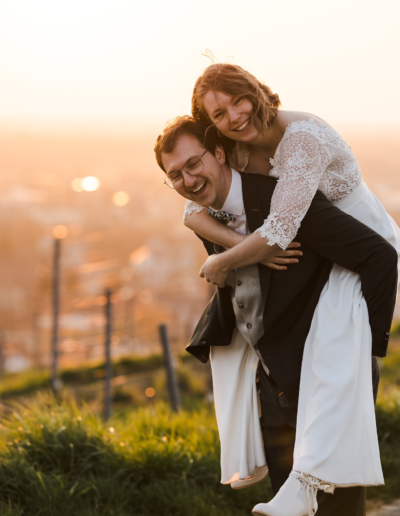 Sunset, die Braut wird huckepack vom Bräutigam getragen. Er lacht mit geschlossen Augen und hat beide Arme um ihre Beine umschlungen. Sie lacht in die Kamera und beide Hände um seine Brust gelegt.