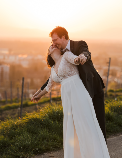 Sonnenuntergang, ein Brautpaar steht auf einer Anhöhe. Sie steht mit dem Rücken an ihn gelehnt und sie halten sich an den Händen, diese sind vom Körper ausgestreckt und sie lachen sich an, neigen sich bei kippend zur Seite.