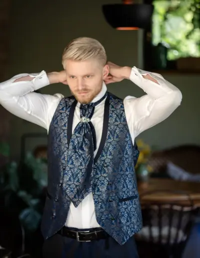 Der Bräutigam richtet sich gerade für die Hochzeit und zieht sich eine Krawatte über. Seine Hände sind hinter seinem Nacken und richten den Kragen. Er lächelt still in sich hinein.