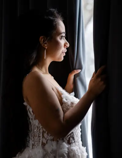Braut steht am Fenster im Raum mit dunklen Vorhängen. Hat beide Hände am Vorhang jeweils links und rechts umklammert und schaut dazwischen nach draussen. Blick ist ernst und nachdenklich.
