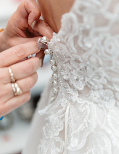 Das Brautkleid wird hinten von der Brautmutter zugemacht. Zu sehen sind Hände und die Knöpfe in Nahaufnahme, sie werden eingefädelt