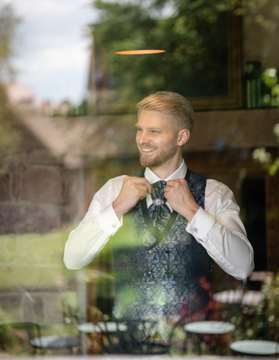 Bräutigam steht hinter einem Glas Fenster im Raum und richtet lachend seine Krawatte, schaut zu Seite, Reflektionen von draussen sind zu sehen