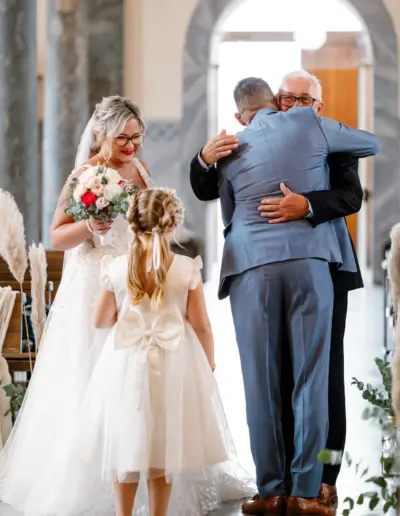 Übergabe der Braut vom Brautvater an den Bräutigam in der Kirche. Der Brautvater umarmt den Bräutigam herzlich und die Braut steht daneben und schaut zu ihrer Tochter.
